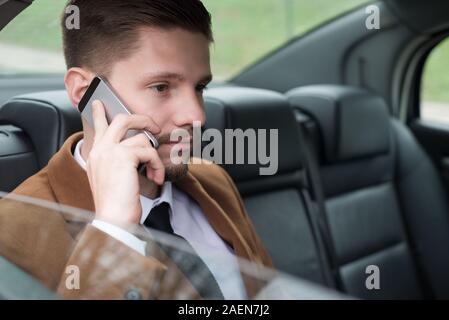 Ritratto di un giovane ragazzo di business. Un imprenditore corse in auto nel sedile del passeggero, parlando al telefono, utilizza uno smartphone. Business man in stylis Foto Stock
