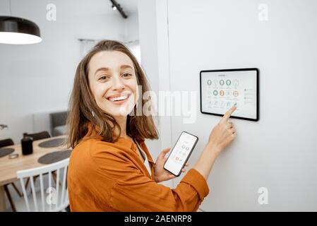 Giovane donna di casa di controllo digitale con pannello touch screen installato sulla parete in soggiorno. Concetto di una casa intelligente e applicazione mobile per la gestione di dispositivi intelligenti a casa Foto Stock