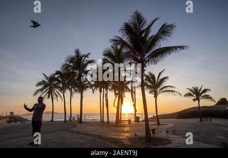 Alba sul Arpoador boulevard con la spiaggia di diavoli e stagliano palme a Rio de Janeiro contro un arancione chiaro e blu cielo Foto Stock