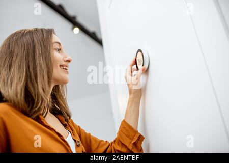 Giovane donna di regolazione temperatura di riscaldamento con un moderno termostato wireless installato sul muro bianco a casa. Smart home regolamento riscaldamento concept Foto Stock