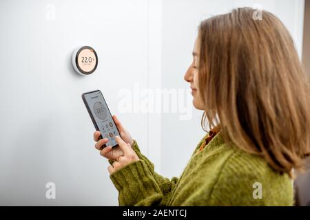 Donna vestita di maglione verde di regolazione temperatura di riscaldamento con un moderno termostato wireless e smart phone a casa. Sincronizzazione del termostato con dispositivi mobili concept Foto Stock