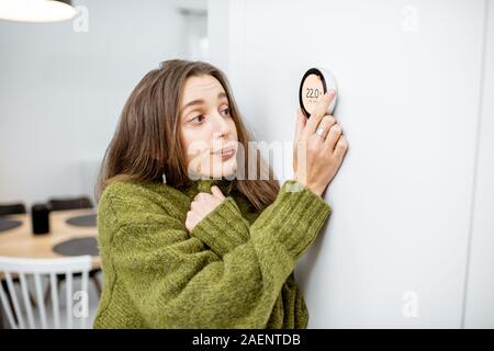 Giovane donna in maglione sensazione di freddo, attesa per la casa a riscaldarsi, controllo impianto di riscaldamento con una smart termostato di casa Foto Stock