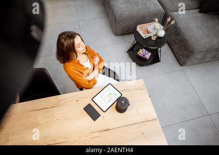 Giovane donna utilizzando dispositivi digitali per smart home control, seduta con tablet e il comando vocale altoparlante a casa. Ampia vista da sopra Foto Stock