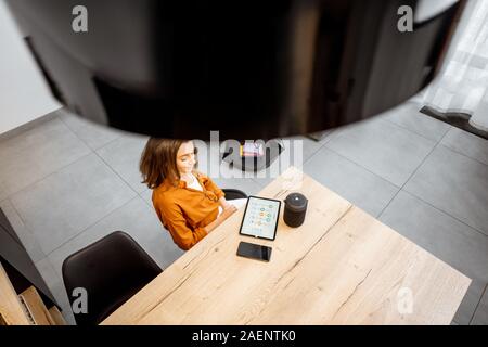 Giovane donna utilizzando dispositivi digitali per smart home control, seduta con tablet e il comando vocale altoparlante a casa. Ampia vista da sopra Foto Stock