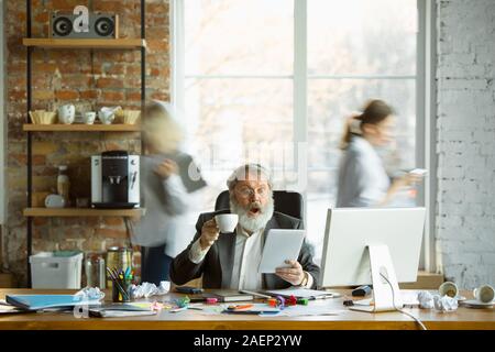 Nervoso e stanco boss al suo posto di lavoro occupato mentre le persone in movimento in prossimità sfocata. Lavoratore di ufficio, manager funzionante, ha dei problemi e scadenza, i suoi colleghi a distrarre. Business, lavoro, concetto del carico di lavoro. Foto Stock