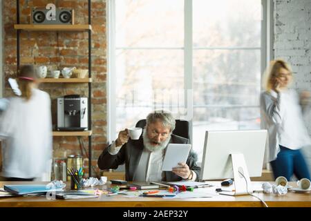 Nervoso e stanco boss al suo posto di lavoro occupato mentre le persone in movimento in prossimità sfocata. Lavoratore di ufficio, manager funzionante, ha dei problemi e scadenza, i suoi colleghi a distrarre. Business, lavoro, concetto del carico di lavoro. Foto Stock