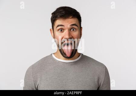 Spensierato divertente e giocoso, Pippo ragazzo con barba, usura maglione grigio, che mostra la linguetta, aprire la bocca e guardare fotocamera, imbrogliamo intorno, mostrando interiore, andare Foto Stock