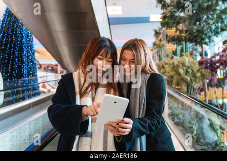 Multi-etnico bella coppia di amiche è di entrare in un grande magazzino sulle scale mobili tramite la digitazione di uno smartphone Foto Stock