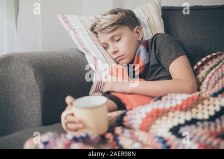 Ragazzo malato a letto con coperta di lana, bottiglie di acqua calda e di una tazza alta. Triste teen con l'influenza si appoggia a casa da solo in una fredda giornata invernale. Bambino con ingredienti di stagione Foto Stock