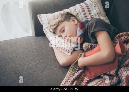 Ragazzo malato a letto con coperta di lana e bottiglie di acqua calda. Triste teen con l'influenza si appoggia a casa da solo in una fredda giornata invernale. Bambino con infettare stagionali Foto Stock