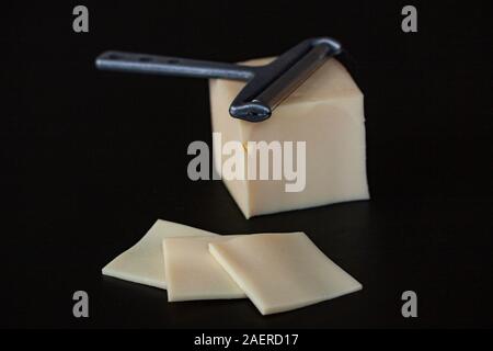 Fette di formaggio giallo e un formaggio slicer giacente su un blocco di formaggio su sfondo nero. Messa a fuoco selettiva chiudere l'immagine. Foto Stock