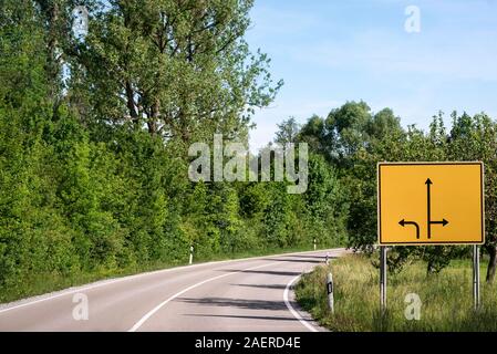 Incrocio un cartello di segnalazione sul lato di una strada vuota in uno scenario primaverile, vicino Obersontheim, Germania. Strada vuota e segno di avvertimento con frecce di direzione Foto Stock