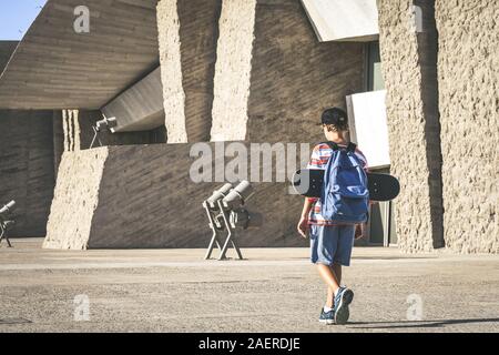 Vista posteriore di un giovane ragazzo con zaino e skateboard in un contesto urbano. Studente alla moda a piedi al di fuori del college dopo un giorno di scuola. La gioventù, nuovo Foto Stock