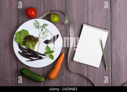 Dieta salutare concetto di piano - appunti per la ricetta, metro a nastro, piastra bianca con foglie di insalata, cetriolo, pomodoro e altri ortaggi sul tavolo di legno Foto Stock