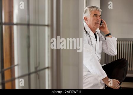 Senior medico maschio siede su un davanzale e utilizza il suo telefono cellulare. Foto Stock