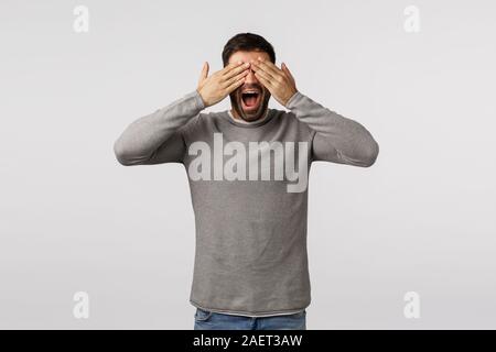 Divertente e spensierata felice bello ragazzo barbuto in grigio maglione, chiudere gli occhi con palme e sorridente si chiedeva, in attesa della bella sorpresa, vogliamo vedere il dono Foto Stock