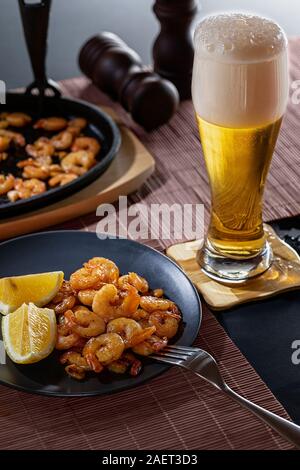 Gamberi fritti e limone in una piastra nera con un bicchiere di birra su sfondo scuro. Close up Foto Stock