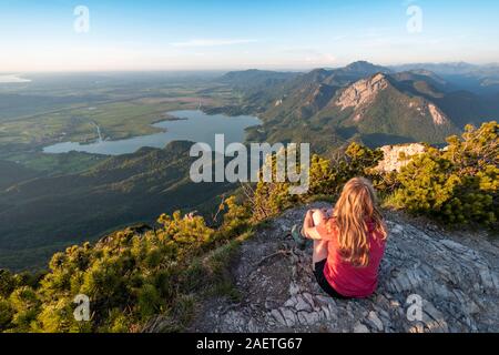 Giovane donna seduta al vertice, vista da Herzogstand al Lago di Kochel, Jochberg e nelle Prealpi Salisburghesi, Alpi Alta Baviera, Baviera, Germania Foto Stock