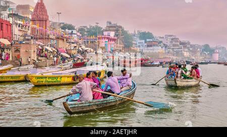 Indian turisti essendo remato lungo il fiume Gange, passando per il lungomare ghats riempito di pellegrini Indù che si bagnano nel fiume santo. Foto Stock