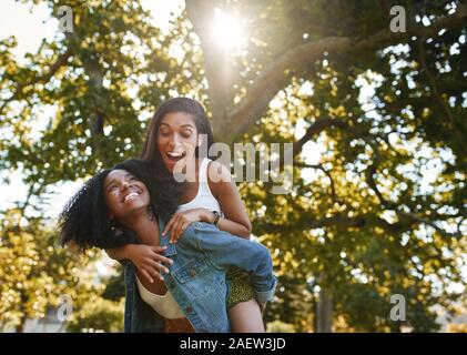 Ritratto di un entusiasta giovane donna godendo i piggyback ride sul suo americano africano amico torna nel parco divertimento e risate Foto Stock