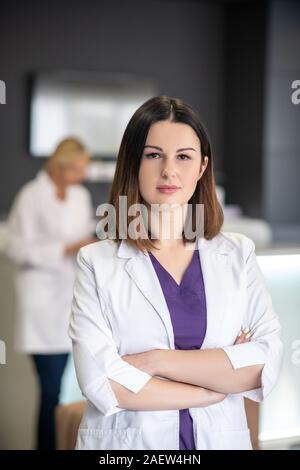 Bella dai capelli scuri medico in veste bianca guardando fiduciosi Foto Stock