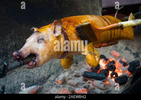 Carcasse di suino vengono grigliate con un'antica stufa a carbone. Il maiale allo spiedo cosparse di olio con un pennello. Arrosto di maiale alla griglia Foto Stock