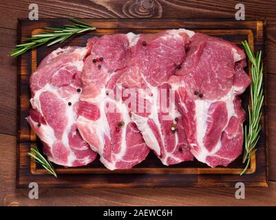 Carne di maiale cruda lombatine su un tagliere con erbe rosmarino al buio su un sfondo di legno, vista dall'alto Foto Stock