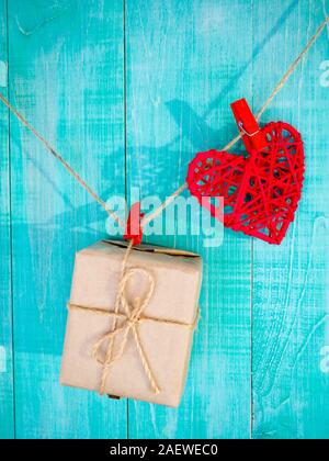 Cuore rosso come simbolo dell'amore il giorno di San Valentino e regalo in carta avvolgente Kraft appeso su abiti su sfondo di legno. Cartolina per il tuo disegno. Foto Stock