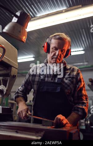 Senior lavoratore manuale nelle cuffie in piedi vicino alla macchina e segare il metallo con SAW Foto Stock
