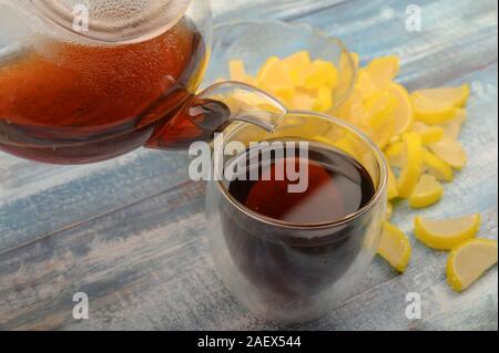 Il tè nero viene versata da una teiera in un bicchiere, marmellata fette di limone su un sfondo di legno. Close up Foto Stock