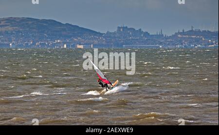 Longniddry Bents, East Lothian, Scozia, Regno Unito. 11 dicembre 2019. Lone windsurf Mikey braves 5 gradi reale sentire -3 temperatura e velocità del vento a 39 km/h SW con raffiche di 58 km/h per godere del suo sport sotto il sole sul Forth Estuary con il castello e la città di Edimburgo e Ponte di Forth Rail e South Queensferry attraversando in background. Foto Stock