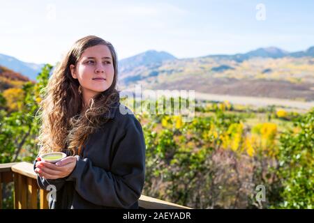 Giovane donna con labbra screpolate in piedi che guarda lontano nel giardino di casa di bere tenendo tazza da tè con vista delle montagne rocciose in Aspen Colorado durante Foto Stock