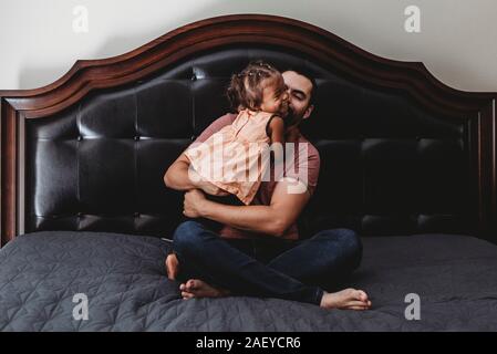 A piedi nudi papà seduta sul letto di ridere con 2 anni figlia biracial Foto Stock