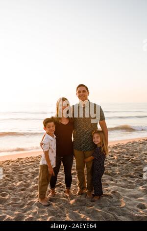 Famiglia di quattro sorridente alla fotocamera, abbracciando sulla spiaggia al tramonto Foto Stock