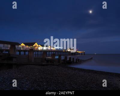 Southwold Pier illuminata di notte accanto a un groyne in legno sotto un chiaro di luna con una nave all'orizzonte, Suffolk Foto Stock