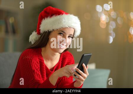 Donna felice tenendo il telefono in natale guardando la telecamera seduta su un divano nella notte a casa Foto Stock