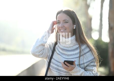Vista frontale il ritratto di una ragazza felice ascolta la musica con gli auricolari wireless e telefono che guarda lontano in un parco Foto Stock