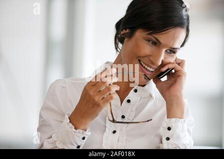 Imprenditrice sorrisi come lei prende una chiamata su un telefono cellulare e tiene i suoi occhiali in mano. Foto Stock
