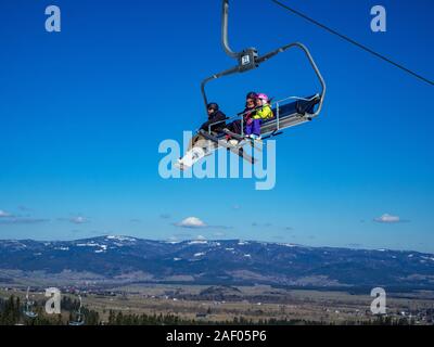 Bialka Tatrzanska, Polonia - 27 Marzo 2018: una seggiovia sul cielo blu con uno snowboarder e sciatori due: una donna e un bambino in un giallo Giacca sci, viole Foto Stock