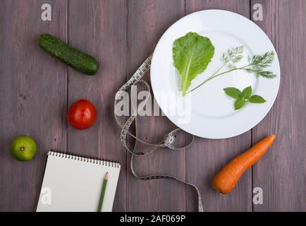 Dieta salutare concetto di piano - appunti per la ricetta, metro a nastro, piastra bianca con foglie di insalata, cetriolo, pomodoro e altri ortaggi sul tavolo di legno Foto Stock