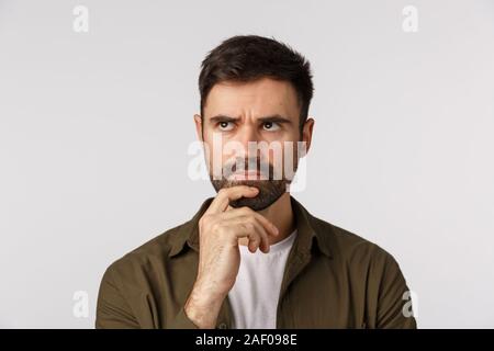 Close-up studio pensieroso barbuto focalizzata giovane uomo in cappotto, toccando il mento e accigliata guardando lontano riflessivo, pensando, considerare cosa, rendendo Foto Stock