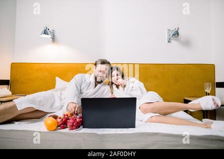 Vista frontale del dolce coppia sposata giacente in letto, guardando il laptop e sorridente. Il marito e la moglie in amore indossando accappatoi bianchi in hotel moderno camera. Foto Stock