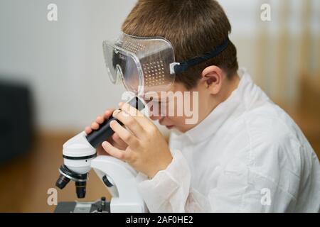 Ragazzo giovane vestito di una tunica bianca e occhiali protettivi osserva attraverso un microscopio in casa sua Foto Stock