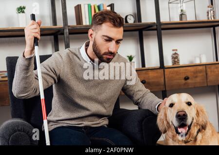 Cieco con bastone seduto in poltrona e accarezzare il golden retriever a casa Foto Stock