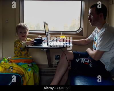 Un giovane padre e un giovane figlio andare su un treno, il Padre guarda il laptop, il bambino gioca macchine da scrivere. La ferrovia, station wagon. Viaggio. Famiglia Foto Stock