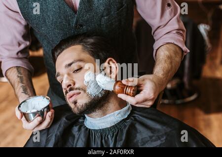 Barbiere di applicare la crema di rasatura sulla faccia del bello uomo barbuto Foto Stock