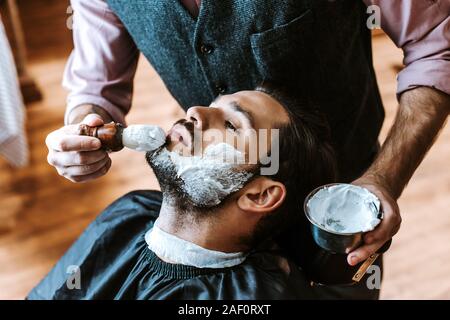 Messa a fuoco selettiva del barbiere di applicare la crema per la rasatura sul volto dell'uomo mentre il recipiente di contenimento Foto Stock