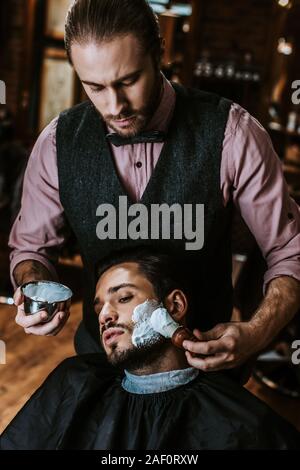 Bel barbiere di applicare la crema per la rasatura sul volto di uomo barbuto Foto Stock
