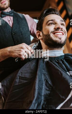 Basso angolo vista del barbiere rifinitore di contenimento mentre per lo styling dei capelli di allegro uomo in Barberia Foto Stock