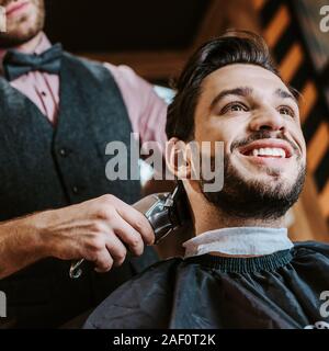 Basso angolo vista del barbiere rifinitore di contenimento mentre per lo styling dei capelli di uomo felice in Barberia Foto Stock